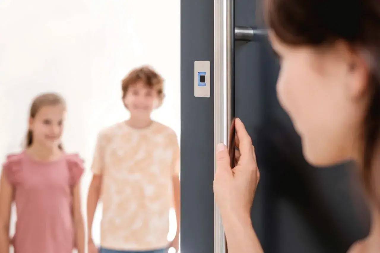 Une personne ouvre la porte d'entrée équipée d'un système de serrure électronique, avec deux enfants visibles en arrière-plan.