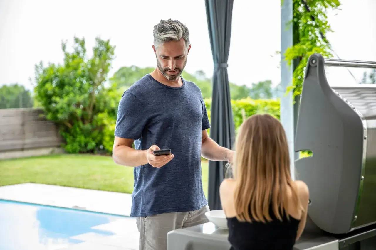 Un homme tenant un smartphone se trouve à côté d'une femme assise près d'un barbecue dans un jardin.