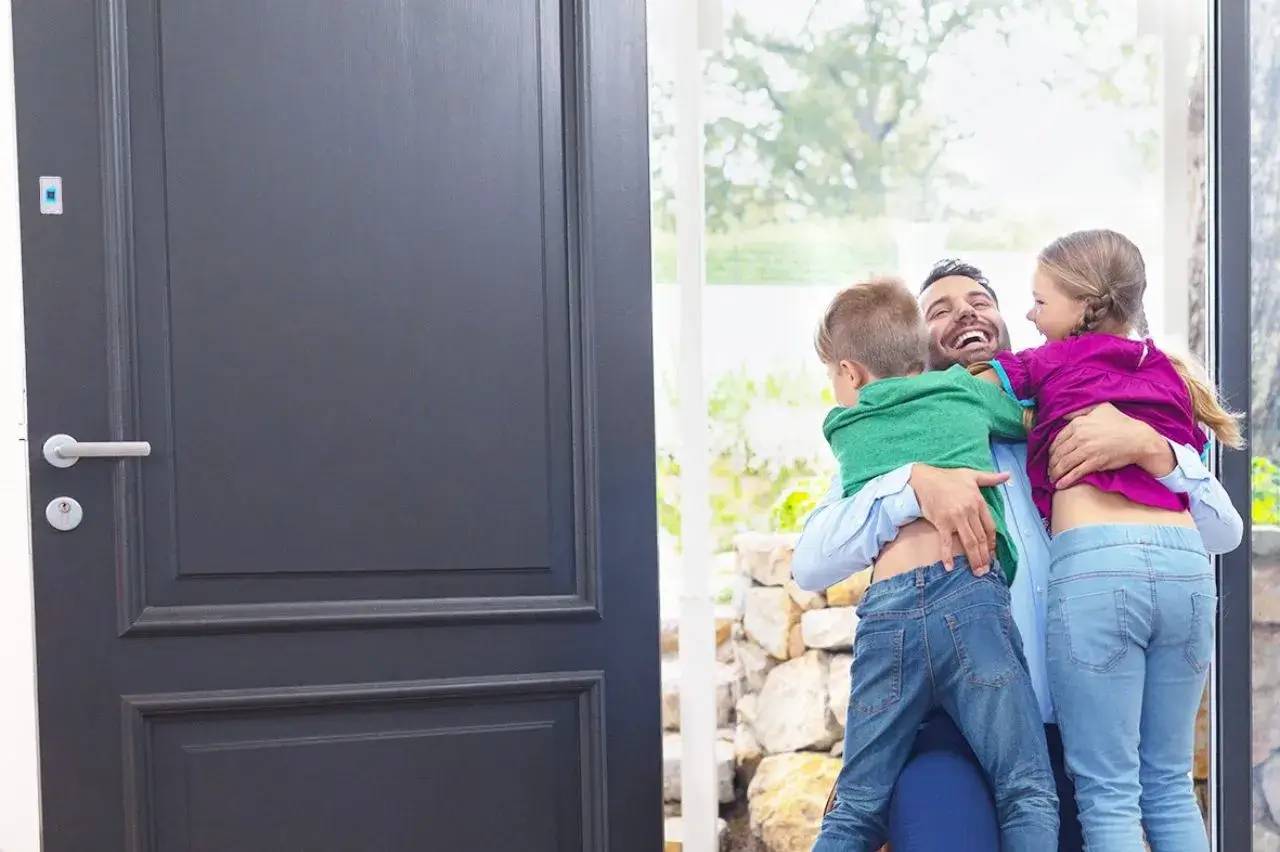 Un homme souriant accueille deux enfants avec un câlin enthousiaste près d'une porte d'entrée.