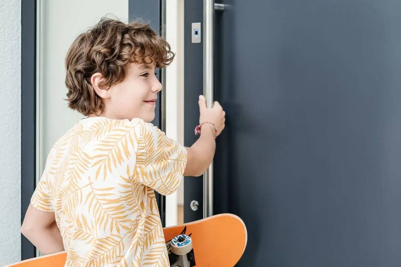 Un enfant souriant, tenant un skateboard orange, ouvre une porte.