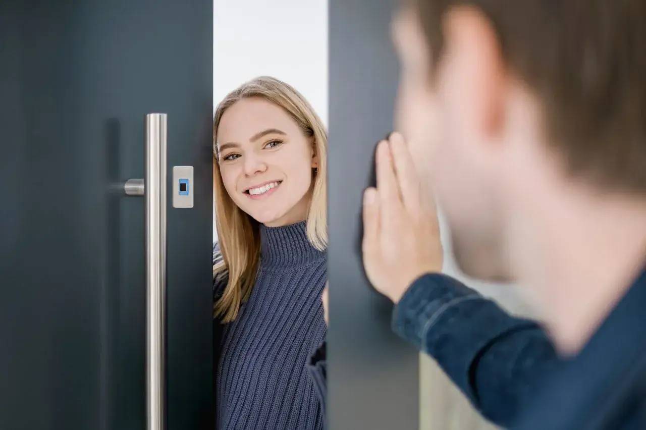 Une femme souriante accueille un visiteur à travers une porte légèrement ouverte.