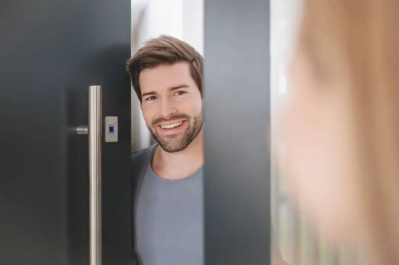 Un homme souriant se tient à une porte entrouverte, donnant une impression d'accueil et de convivialité.