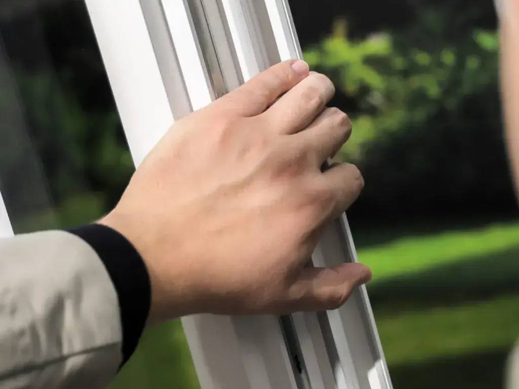 Une main tient le cadre d'une fenêtre ouverte, avec un fond de verdure floue à l'extérieur.