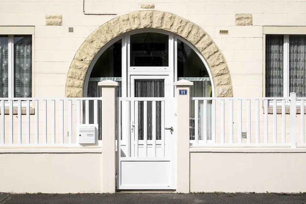 Façade de maison beige avec une porte blanche entourée d’un arc en pierre et une clôture assortie.