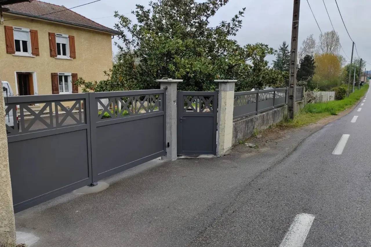Une maison beige avec des volets en bois est bordée par un portail gris moderne le long d'une route.