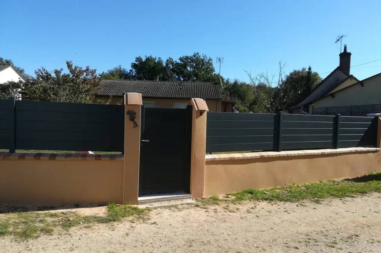 Un portail moderne en métal gris encadré par un mur beige sépare une maison et son jardin de la rue.