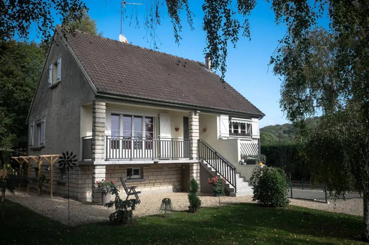 Maison traditionnelle à un étage avec jardin arboré, montée d'escaliers et terrasse en façade, sous un ciel bleu.