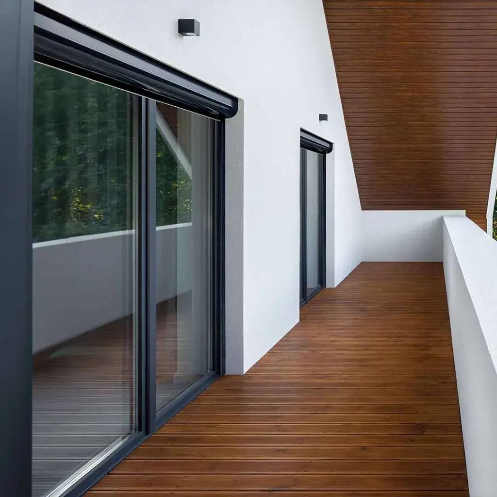 Balcon moderne avec un sol en bois et grandes baies vitrées encadrées de noir, offrant une vue sur la nature environnante.