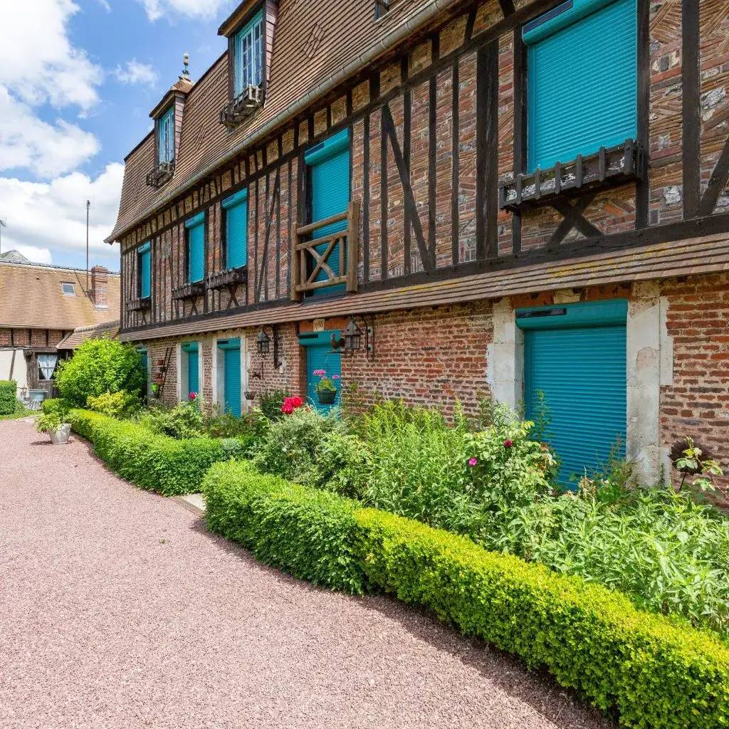 Cette image montre une maison à colombages avec des volets bleus vibrants, entourée de verdure et de fleurs éclatantes sous un