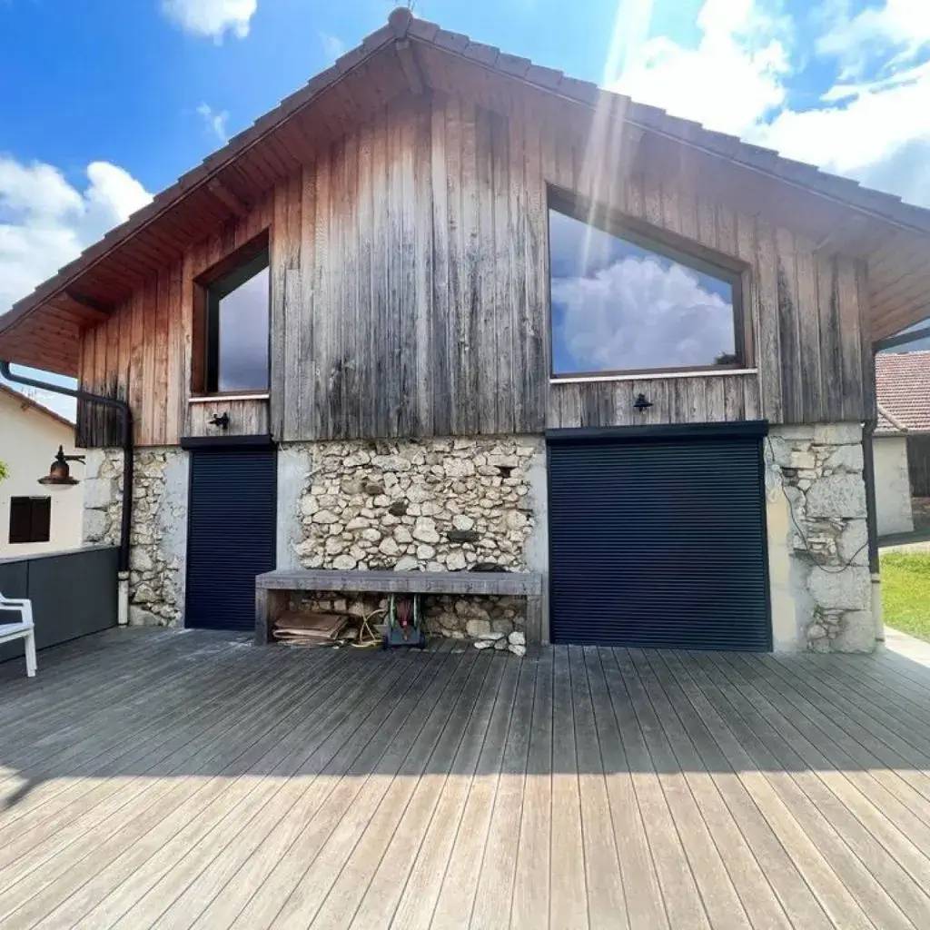 Un bâtiment en bois avec une façade en pierre, doté de grandes fenêtres triangulaires et de volets roulants, sous un ciel bleu avec quelques