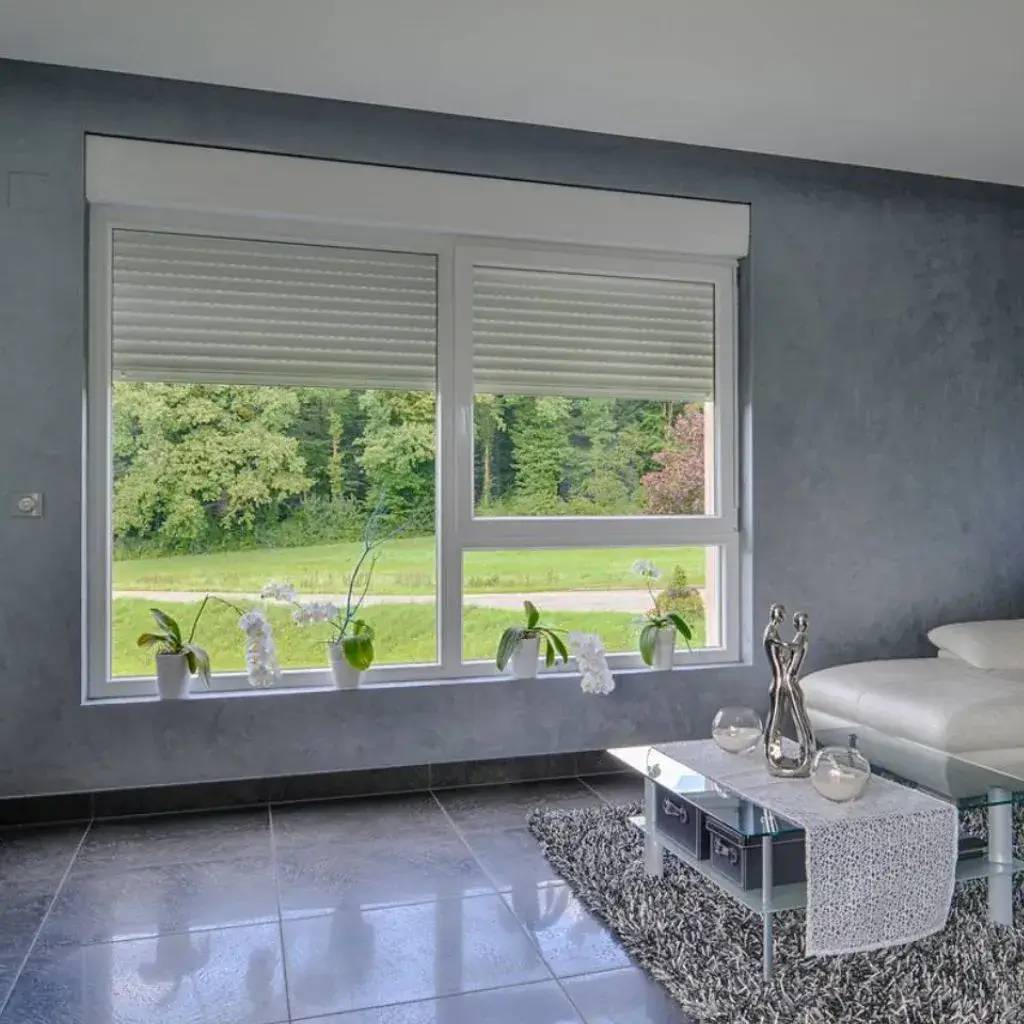 Une pièce moderne avec un sol carrelé et un grand canapé blanc, donnant sur un jardin verdoyant à travers une grande fenêtre équipée de stores en