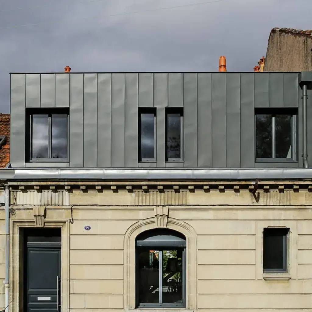 Une façade de bâtiment alliant modernité et tradition, avec un revêtement gris métallique sur le toit et une base en pierre beige.