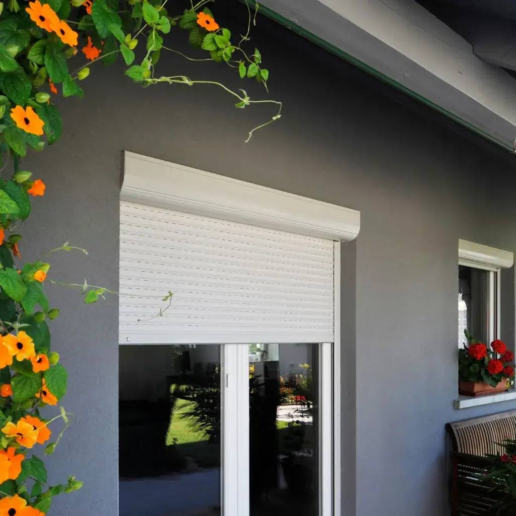 Une fenêtre moderne avec volet roulant blanc sur une façade grise, ornée de fleurs grimpantes orange et d'un bac à fleurs rouges.