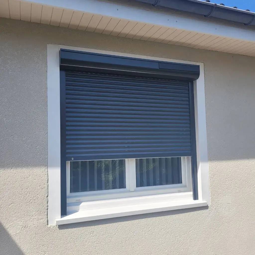 Une fenêtre moderne avec un volet roulant gris foncé partiellement abaissé sur une façade beige.