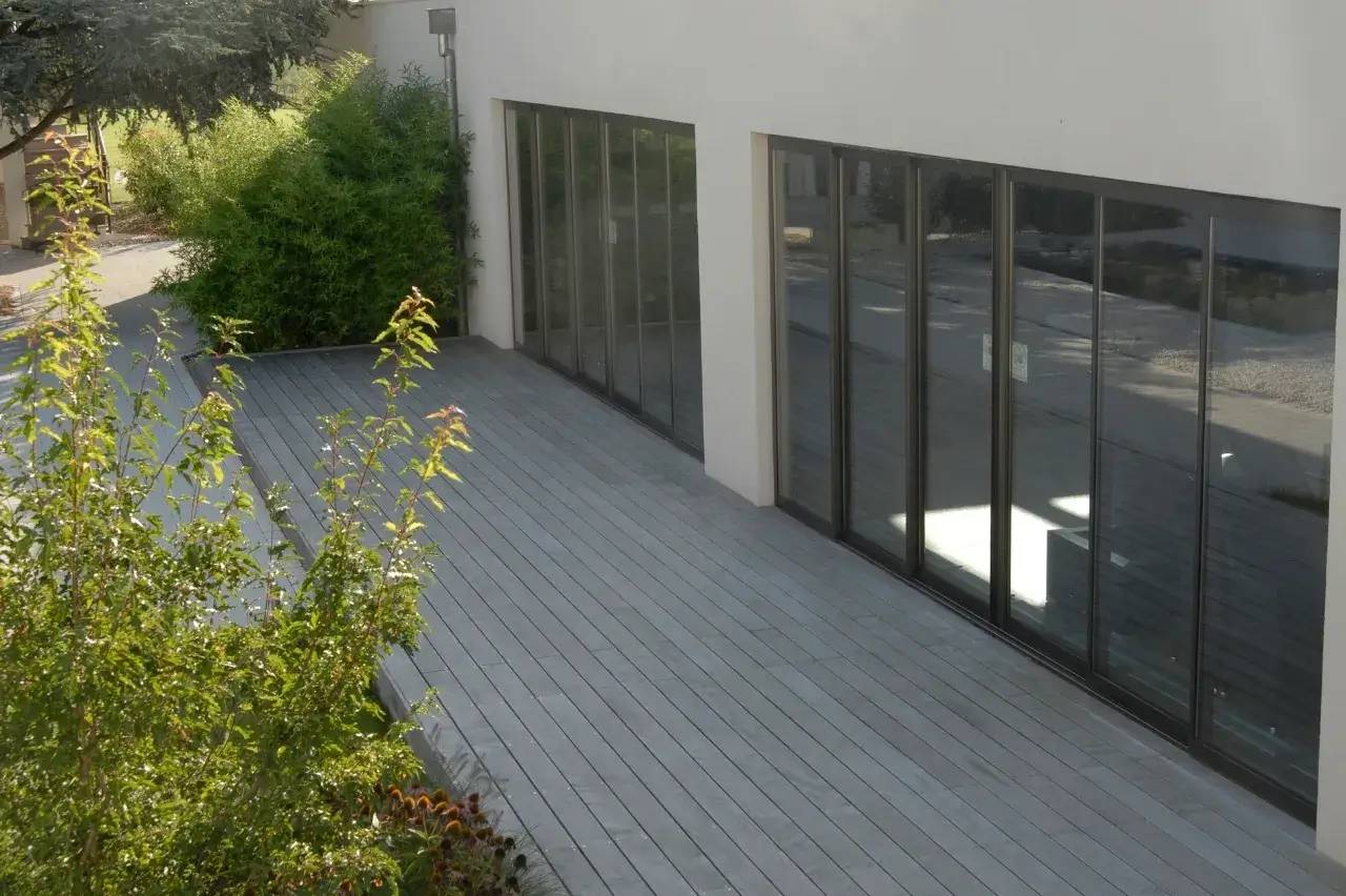 Une terrasse en bois gris longe une façade moderne avec de grandes baies vitrées, entourée de verdure.