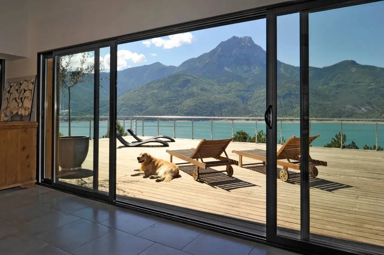 Vue panoramique d'une terrasse ensoleillée avec des chaises longues, un chien et un paysage de montagne et de lac à travers de grandes ba