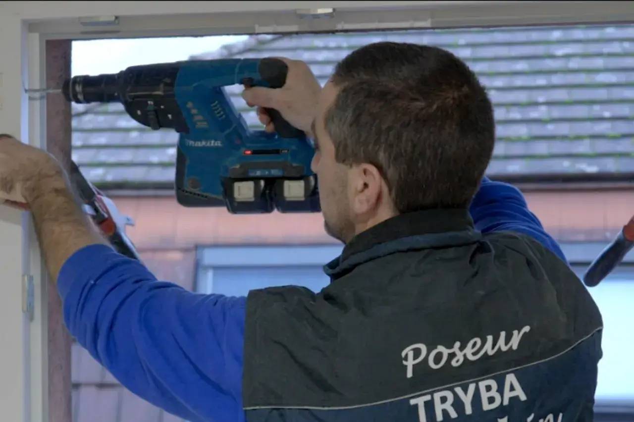 Un poseur de fenêtres utilise une perceuse pour installer un cadre, vêtu d'une veste avec le logo Tryba.