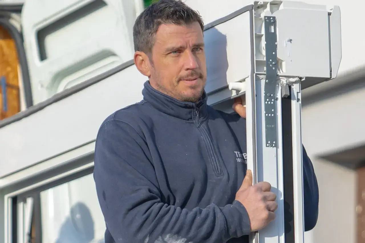 Un homme en tenue de travail tient un grand cadre métallique devant un bâtiment.