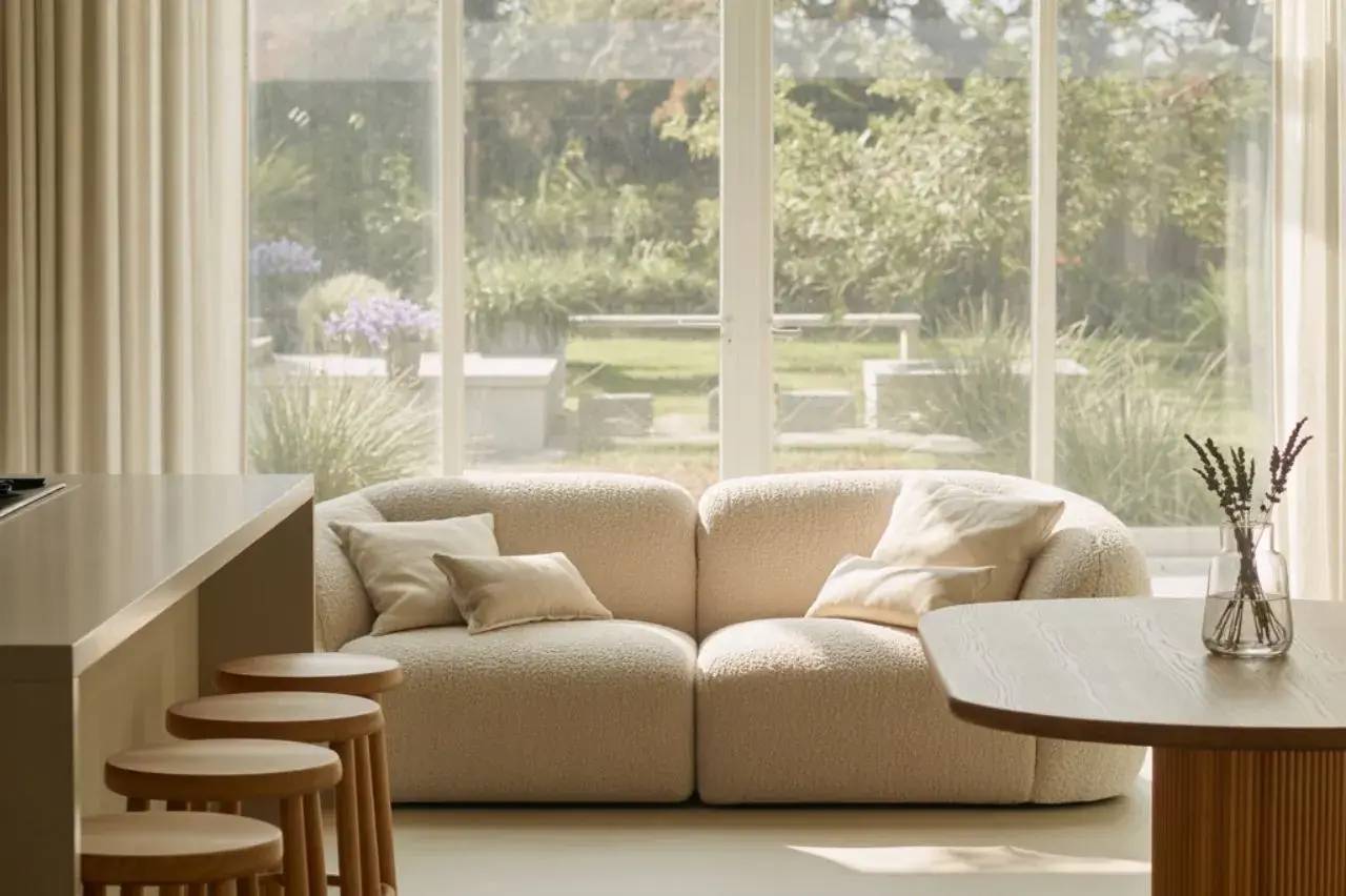 Un salon lumineux avec un canapé beige, des coussins assortis, et une vue dégagée sur un jardin verdoyant.