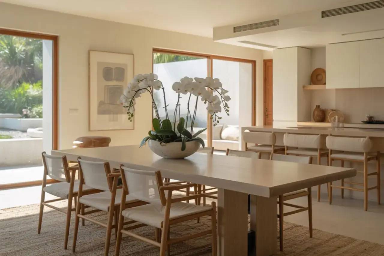 Salle à manger élégante avec table en bois clair et orchidées blanches, baignée de lumière naturelle.