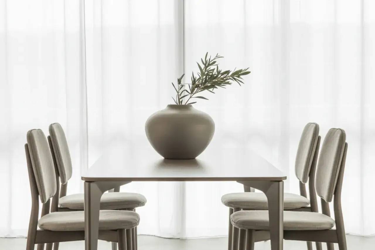 Une table minimaliste en bois clair est entourée de quatre chaises, avec un vase en céramique et une branche de feuillage au centre