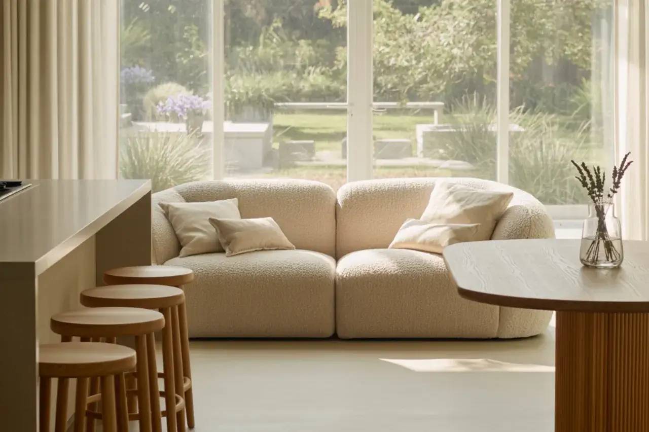 Un salon lumineux avec un canapé beige confortable, entouré de tabourets en bois et offrant une vue sur un jardin verdoyant.