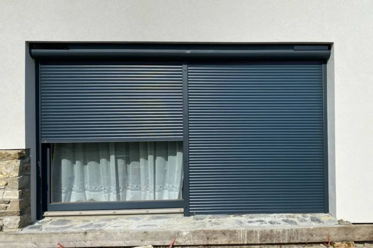 Un volet roulant gris foncé enveloppe partiellement une fenêtre avec un rideau blanc derrière, sur fond de mur clair.