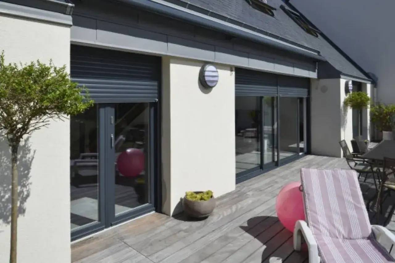 Une terrasse en bois avec des chaises longues, des fenêtres à volets roulants et des pots de plantes, offrant un espace extérieur moderne et accueill