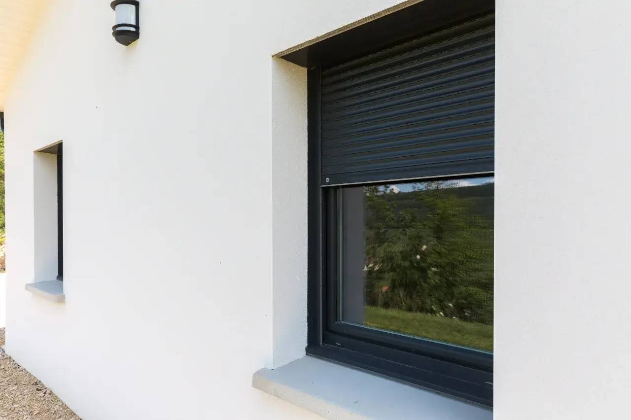 Fenêtre moderne avec volet roulant noir sur une façade blanche, éclairée par une applique murale.