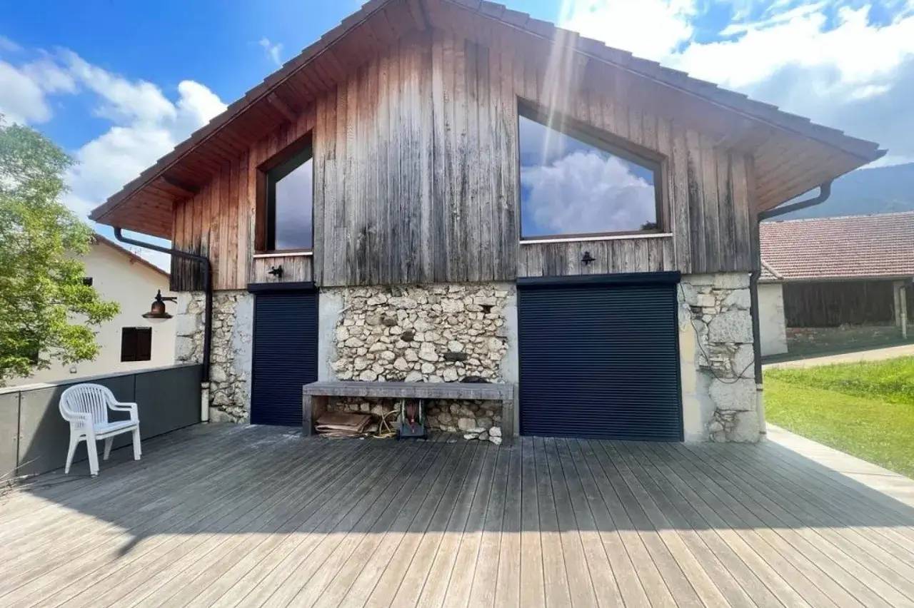 Un chalet en bois moderne avec des murs en pierre, doté de grandes fenêtres et d'une terrasse en bois, baigné de lumière naturelle sous un ciel