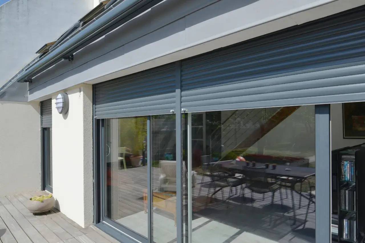 Des volets roulants gris foncé couvrent les grandes baies vitrées d'une maison moderne, laissant entrevoir une terrasse en bois et un