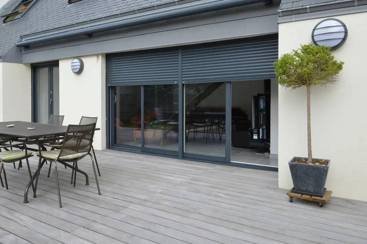 Une terrasse en bois avec un mobilier de jardin moderne, des portes coulissantes vitrées et un arbre en pot élégant près de la façade.