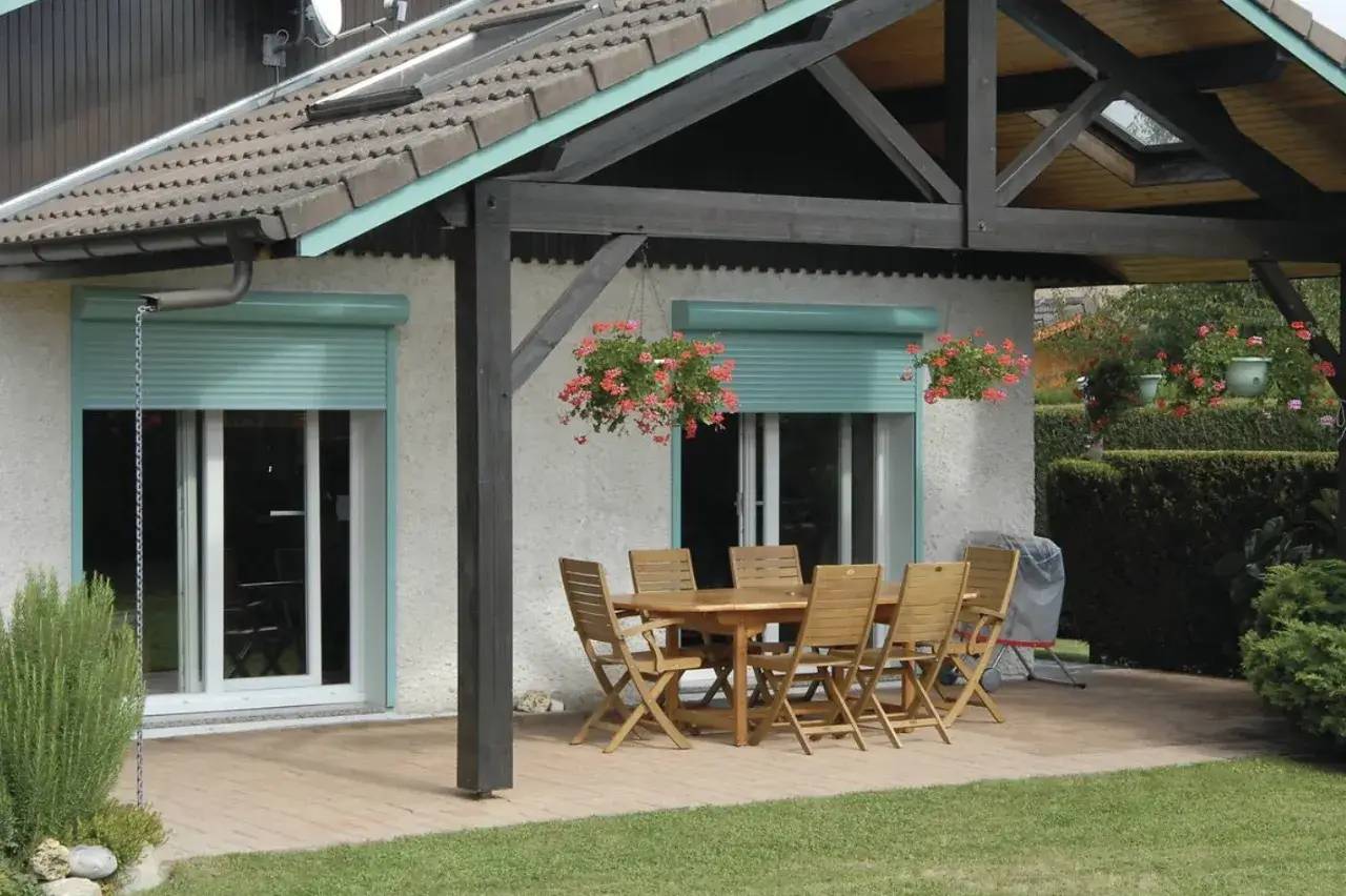 Une terrasse en bois avec une table et des chaises en plein air, protégée par une pergola, entourée de verdure.