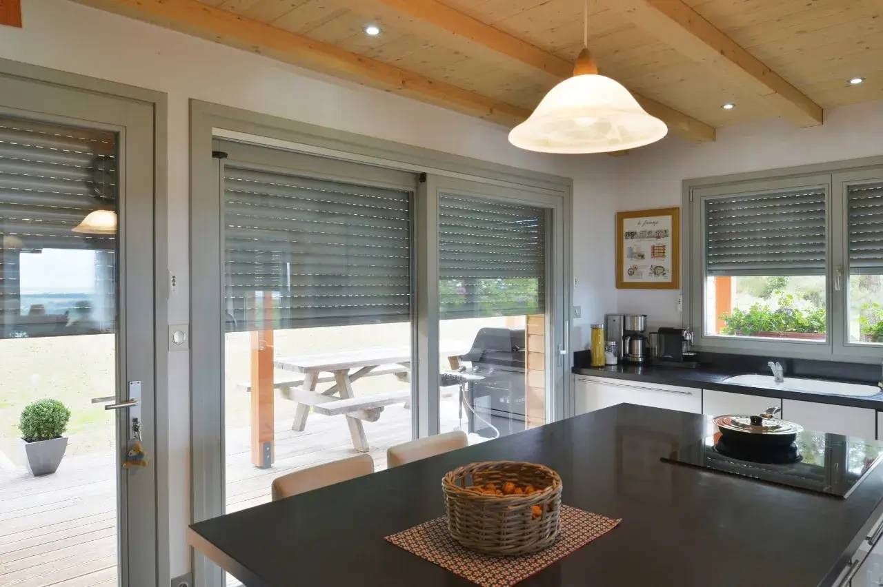 Cuisine moderne avec des volets roulants, un îlot central avec un panier de fruits et une vue sur la terrasse extérieure.