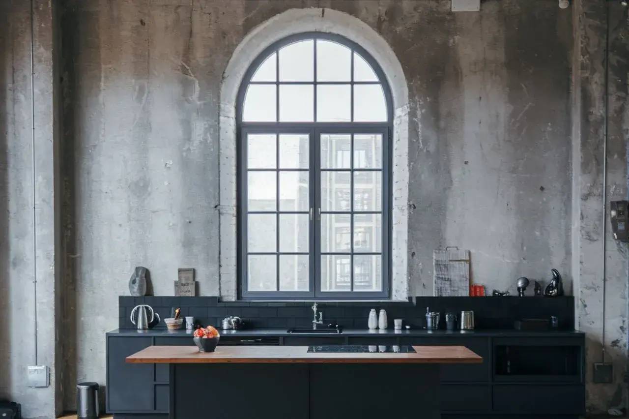 Une cuisine industrielle épurée avec des murs en béton brut, un grand plan de travail noir et une fenêtre arche encadrée de fer.