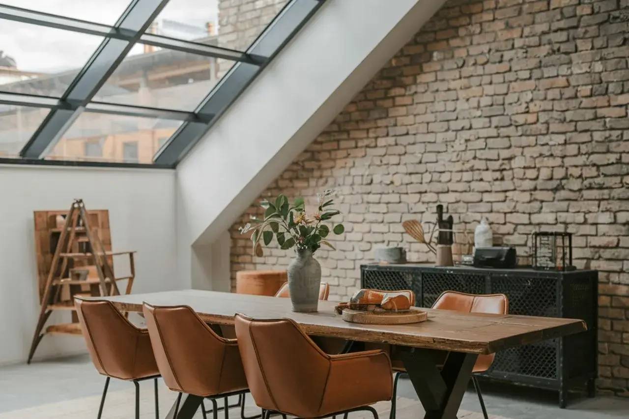 Un espace de salle à manger moderne avec table en bois, chaises en cuir marron, et mur de briques apparentes sous un puits