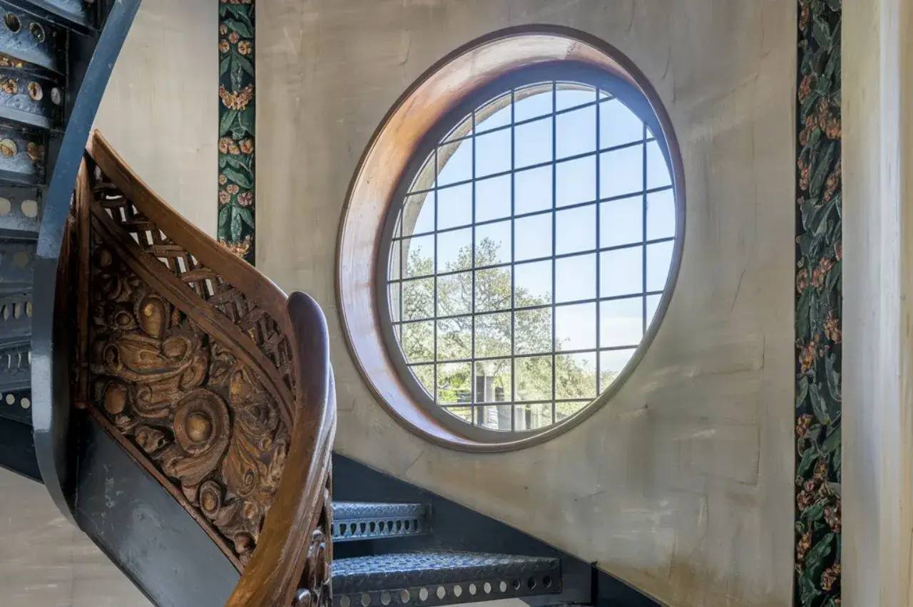 Un escalier hélicoïdal en métal et bois sculpté mène à une grande fenêtre ronde avec vue sur des arbres.