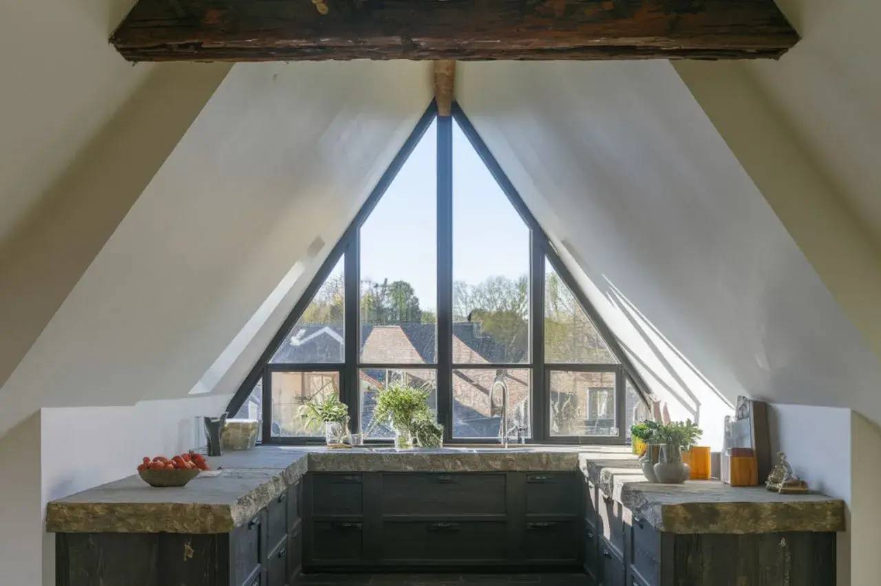 Une cuisine moderne et lumineuse avec une grande fenêtre triangulaire offrant une vue sur l’extérieur et des plans de travail en pierre.