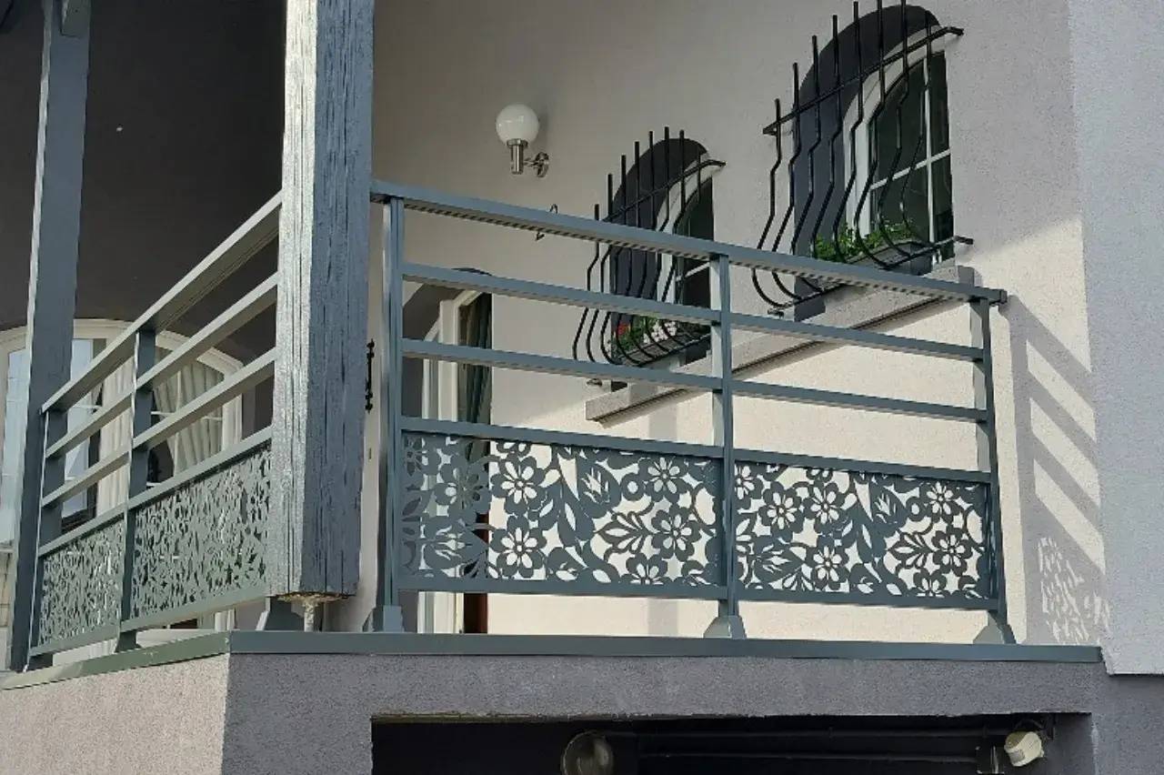 Un balcon moderne de couleur grise avec une rambarde ornée de motifs floraux en métal découpé, surplombe une façade claire dot