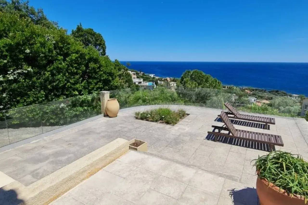 Une terrasse ensoleillée surplombe la mer, entourée de verdure, avec des chaises longues en bois et un grand pot en terre