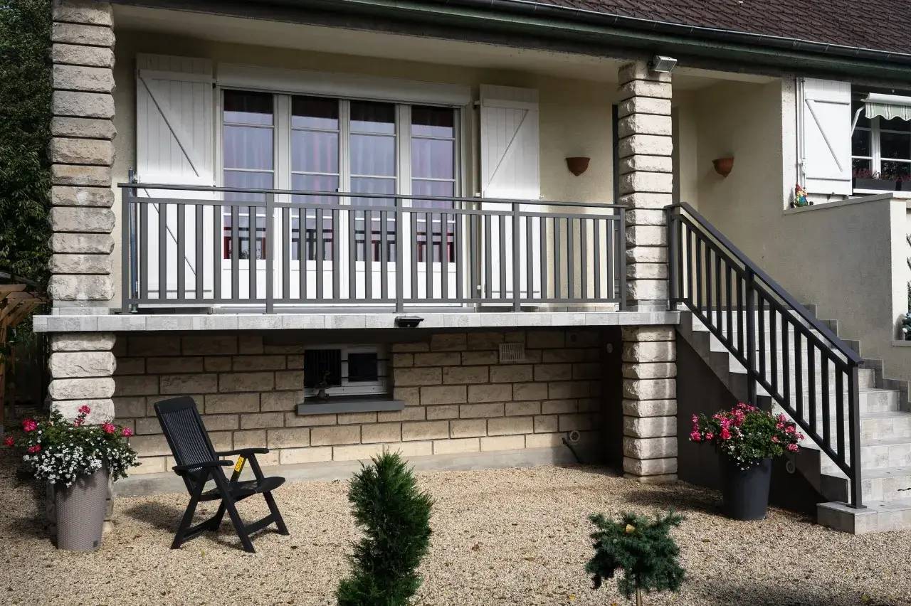 Façade de maison avec balcon en bois gris, escalier, et jardin gravillonné agrémenté de plantes en pot.