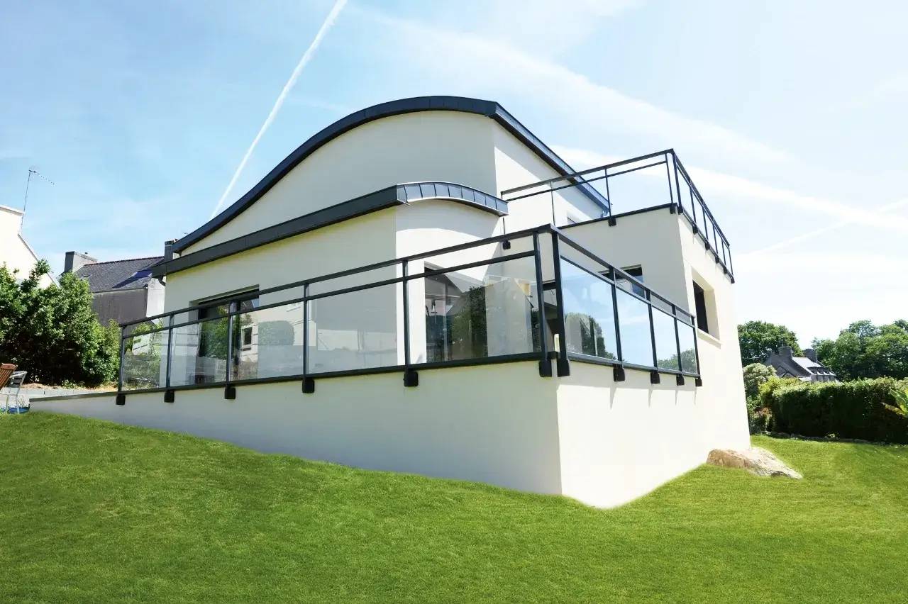 Maison moderne à toit ondulé avec une terrasse en verre surplombant une pelouse verdoyante.