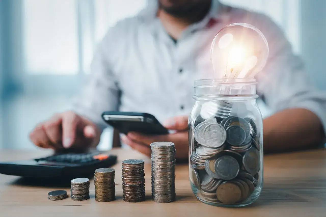 Un homme utilise une calculatrice à côté de piles de pièces et d'un bocal rempli de monnaie surmonté d'une ampoule allumée