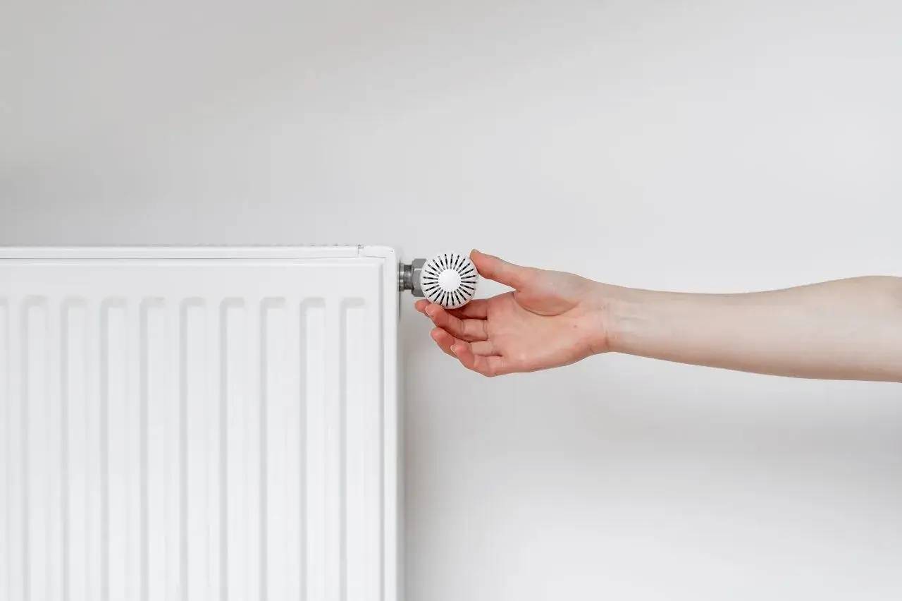 Une main ajuste la vanne thermostatique d'un radiateur blanc sur fond clair.
