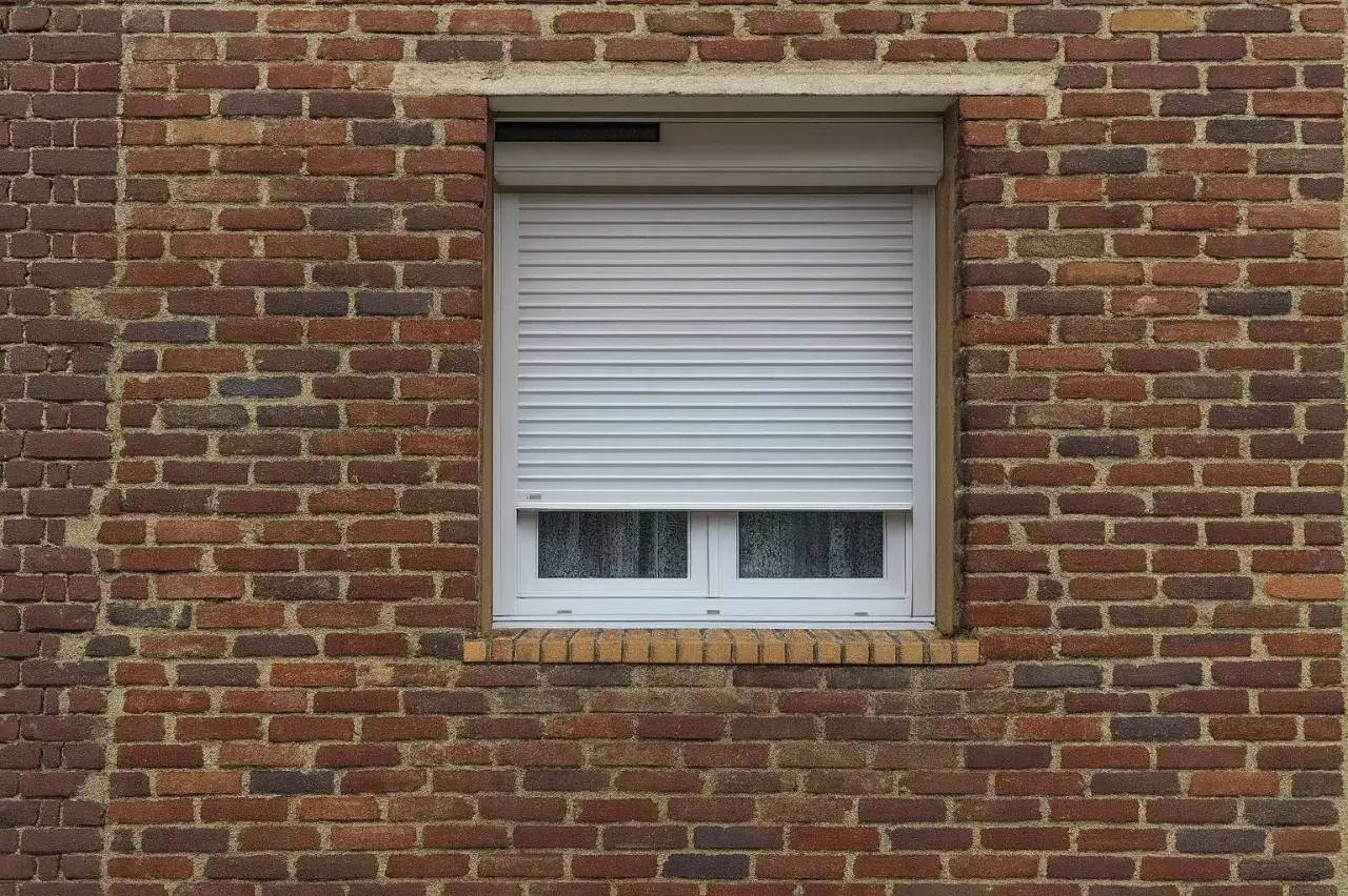 Fenêtre avec volet roulant blanc sur un mur de briques rouges.