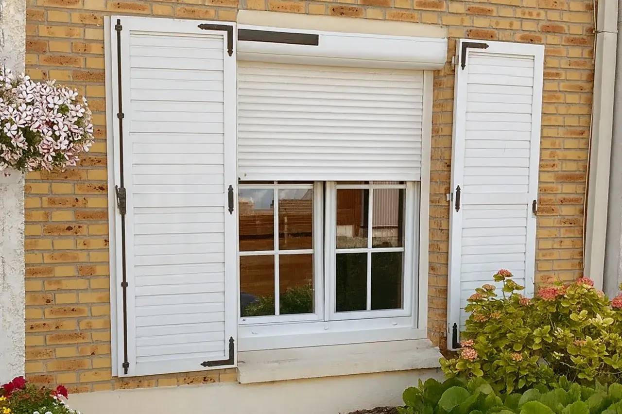 Fenêtre en PVC avec volets roulants et battants, encadrée de briques, entourée de fleurs et de verdure.