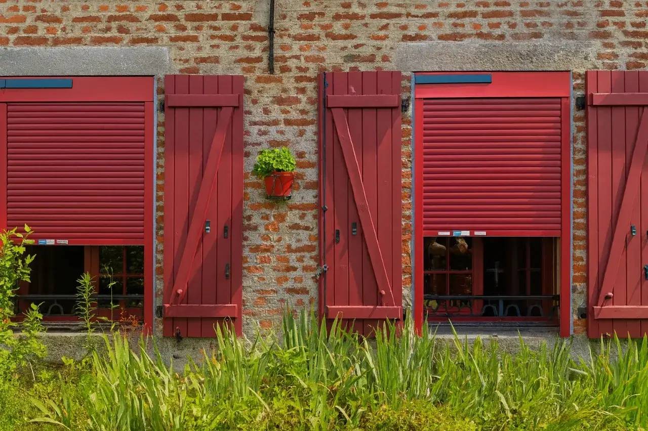 Façade en briques avec trois volets rouges fermés, dont un muni d'un volet roulant partiellement abaissé, et un pot de