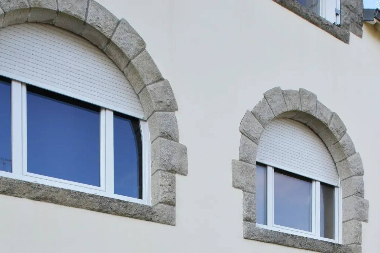 Deux fenêtres arquées avec volets en pierre grise et cadre blanc se détachent sur une façade beige.