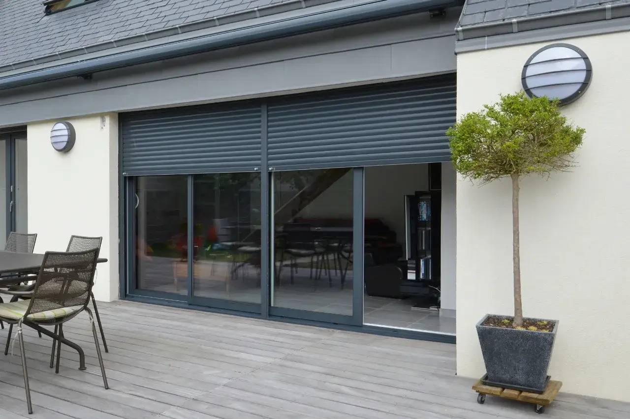 Terrasse en bois moderne avec fauteuils et table, donnant sur de grandes baies vitrées équipées de volets roulants.