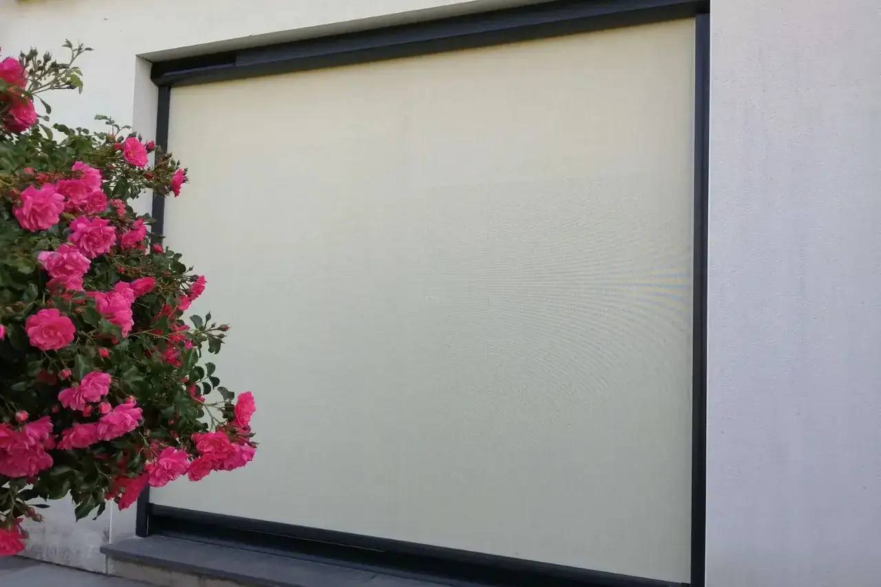 Un mur blanc est partiellement couvert par un volet roulant beige, avec des fleurs roses en premier plan sur la gauche.